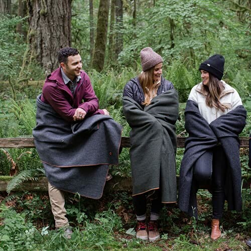 Three people sitting on a fence in a forest, wrapped in blankets.