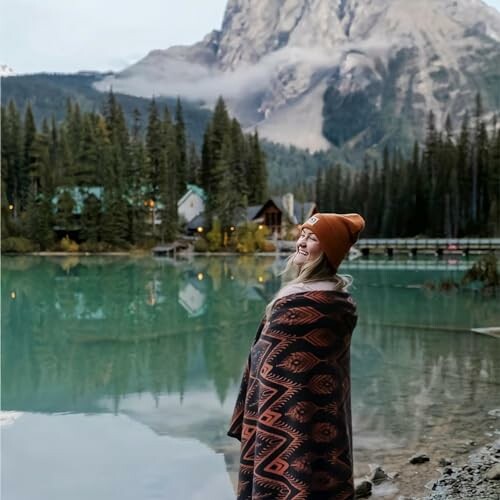 Woman in beanie and blanket by a mountain lake.